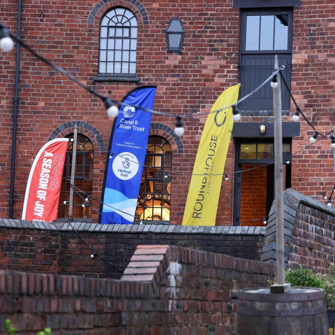 A square image showing a bricked building and courtyard. At the back of the image is three banners. From left to right, these banners include: a small red banner, a blue banner with a circular logo in the middle and a yellow banner with a smiley face logo at the top. Behind these banners is a brick building, and there are arched windows and a open doorway. In the foreground of the image is festoon lighting. 