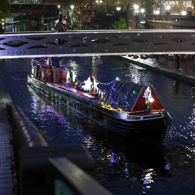 A square image showing a heritage working boat on a canal. The boat is covered in fairy lights and other lanterns. In the boat is a flautist and a drummer. In the background is streetlights, and across the canal is a bridge.