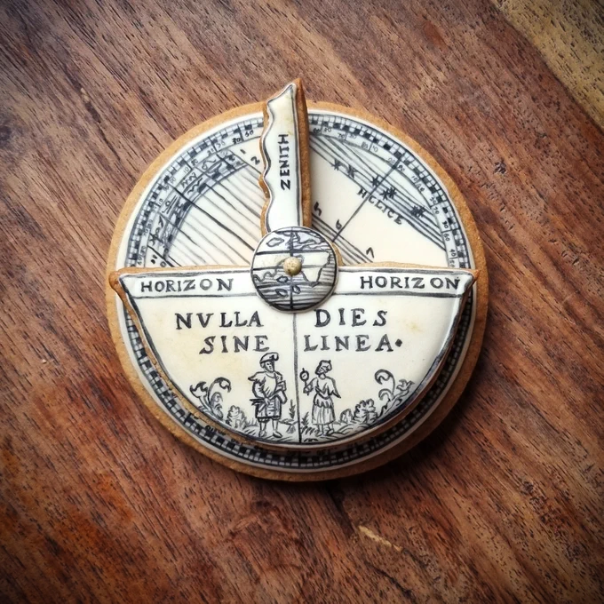 Two biscuits stacked on top of one another, designed to look like a rotating diagram from an early modern astronomical book. The lower biscuit is circular and features a series of horizontal lines and tiny numbers in an early modern font. The top biscuit is roughly in the shape of boat: the 'mast' area reads 'zenith'; the main body of the shape features tiny figures in historical dress holding astronomical instruments and a Latin motto reading 'Nulla dies sine linea'.