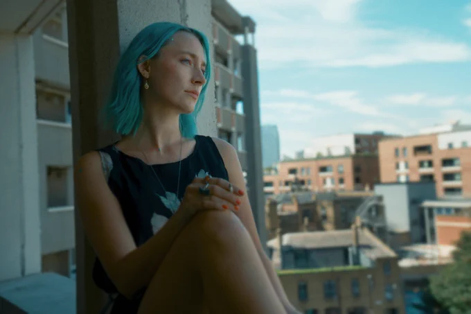 A film still from The Outrun featuring Saoirse Ronan with blue hair, sat on the balcony edge of a tower block. More buildings can be see on the distance. 