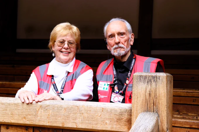 Two volunteer stewards sit in the gallery of the Globe Theatre. They both smile at the camera. 