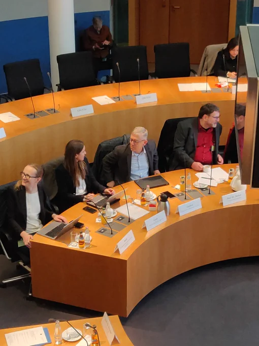 Peter Ganten sitzt im Ausschusssaal an seinem Tisch, rechts und links neben ihm sitzen andere Sachverständige (Stefan Decker, Adriana Groh, Bianca Kastl): Peter Ganten ist Adriana Groh zugewandt, die links neben ihm sitzt, und ist mit ihr im Gespräch.