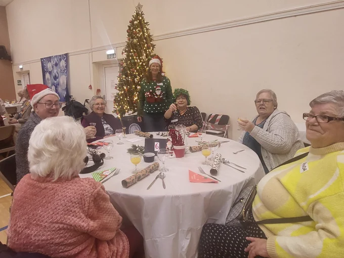 Clients sit around a table. A volunteer is ready to serves drinks. There is a Christmas tree.