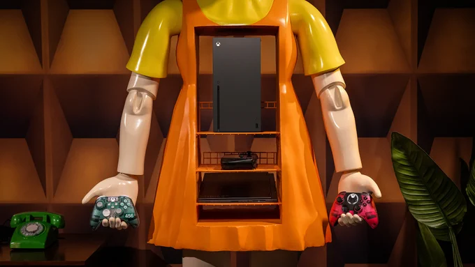 Close up image of the Squid Game Young Hee Gaming Cabinet holding the Squid Game Game Over Controller in the left hand and holding the Squid Game Pink Guard controller in the right hand. Inside the cabinet is an Xbox Series X, a ROG Ally X Black Handheld Gaming Device, and a Lenovo Legion Slim 5 Gen 8.