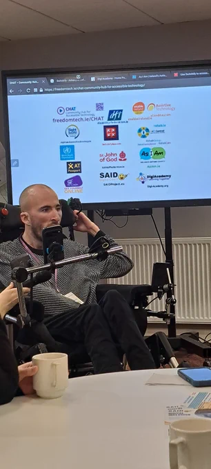 Owen is presenting using his mobile phone and a head swutch, in front of a screen with logos of the stakeholders involved in CHAT.