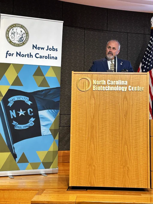 A person stands at a podium with the North Carolina Biotechnology Center logo, speaking at an event about "New Jobs for North Carolina" with state flags in the background.