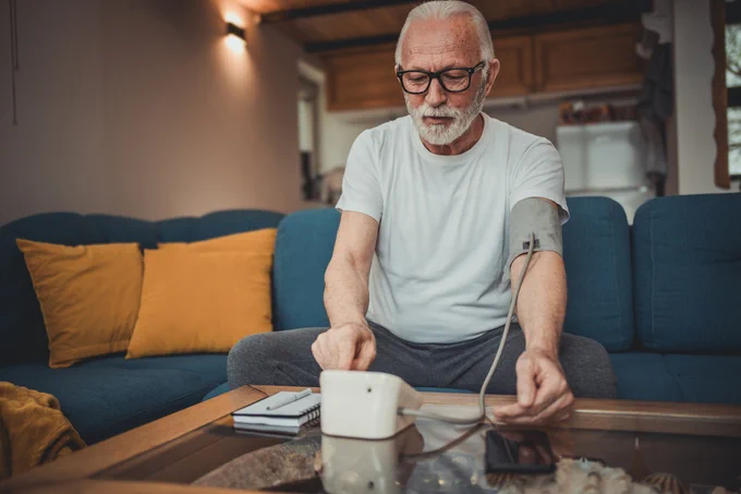 An elderly man dressed in a white t-shirt and grey sweatpants is seated on a blue sofa, using a blood pressure monitor and looking at a smartphone in a cozy living room setting.