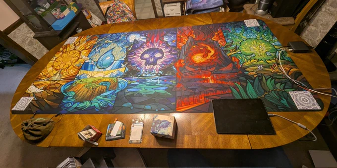 An over table shot of a full table length playmat. The art is of all five of Magali Villeneuve stained glass land arts.