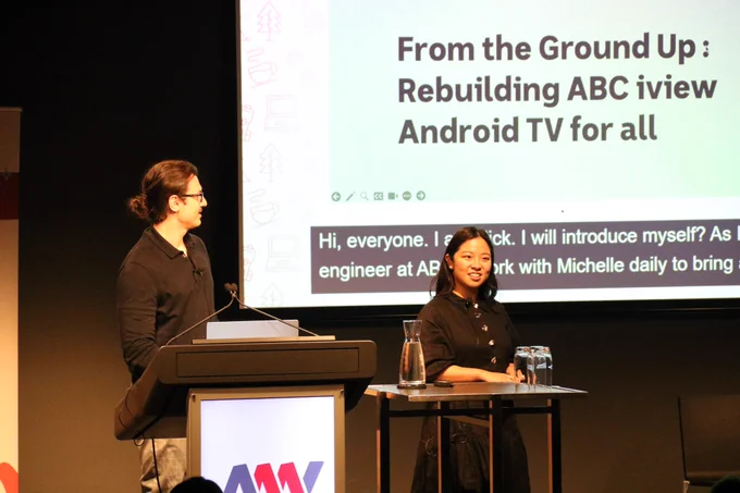 Michelle Chu and Nick Stathakis at A11y Camp, presenting at a lectern in front of a big screen, which shows their slides and the live captions.