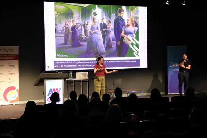 Maia Miller presenting on stage at A11y Camp. There is a large photo of the Bridgerton cast all dressed in purple on the screen behind her. An Auslan interpreter is on the right of stage.