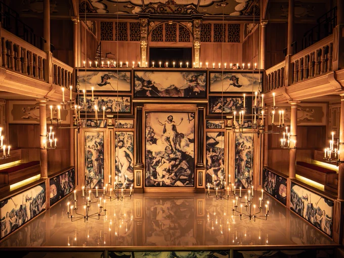 The set for All's Well that Ends Well (2024/5). 

Interior of the Sam Wanamaker Playhouse decorated with with murals of the Last Judgement. Multiple lit chandeliers illuminate the playhouse. The light from the candles reflects on the shiny white surface of the stage. 