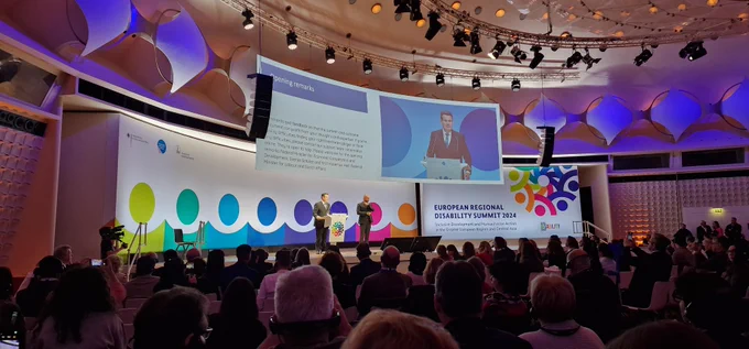 German Federal Minister for Labour and Social Affairs opens the Summit from a stage in Berlin. Backdrop shows colourful logo of the European Regional Disability Summit, a large screen with simultaneous captioning, and there is an audience in front of the state.