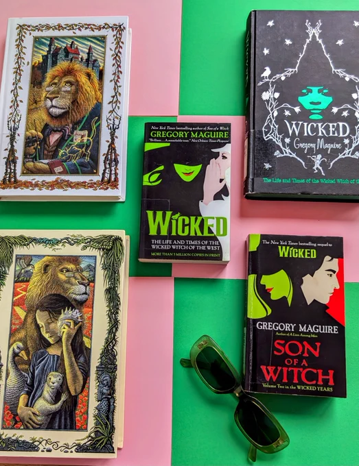 A flatlay of 5 books from the "Wicked" series on a green and pink background with a pair of green sunglasses 