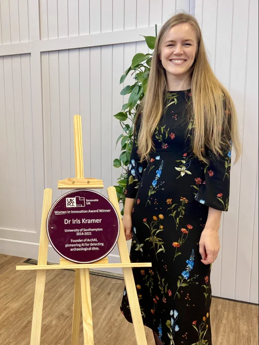 Dr Iris Kramer with the purple plaque: Dr Iris Kramer, a young woman with long blonde hair, stands smiling beside a purple plaque mounted on a wooden easel. She wears a black floral dress, and the plaque highlights her achievements as the founder of ArchAI, pioneering AI for detecting archaeological sites.
