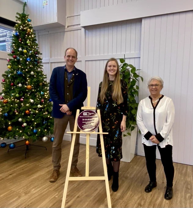 Dr Iris Kramer with Prof Tim Norman and Lou Williams: Dr Iris Kramer stands between Prof Tim Norman and Lou Williams in a brightly lit room. A Christmas tree adorned with colorful ornaments stands in the background. Dr Kramer smiles beside her purple plaque, which is mounted on a wooden easel.