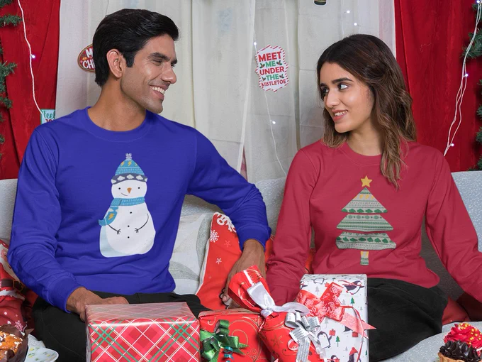 A man and a woman sit on a sofa, surrounded by cakes and wrapped gifts. The man wears a sweater with a snowman design on it. The Snowman has clouds on the rim of his hat. The lady wears a sweater with a Christmas tree on, which has cloud shaped decorations going around it.