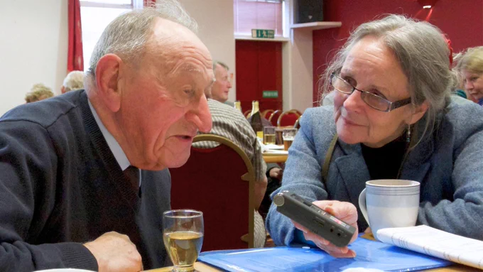 Photo of a woman in a blue blazer and glasses holding out a tape recorded to an older man seated at a table