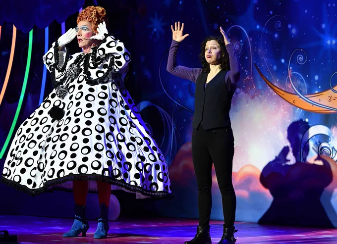 Two peopleon stage during a theatrical production, one dressed elaborately in a black and white polka dot costume and the BSL interpreter is in a simple black outfit, in front of a whimsical, starry backdrop.

