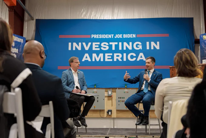 ALT: Secretary Pete Buttigieg and Mayor Mueller sit in a fireside chat. The sign behind them reads: President Joe Biden. Investing In America 