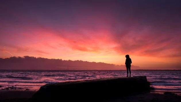 A silhouette stands on a long flat rock by the ocean looking out at a yellow, pink and purple sunset