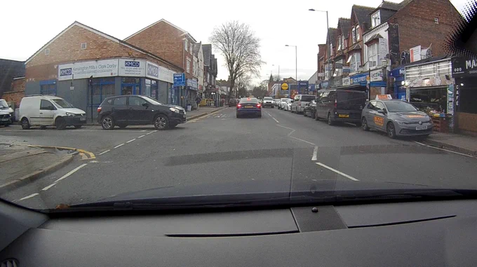A grey Volkswagen with Bill Plant Driving School livery is parked partially on the footway, on white zigzags approaching a pedestrian crossing.