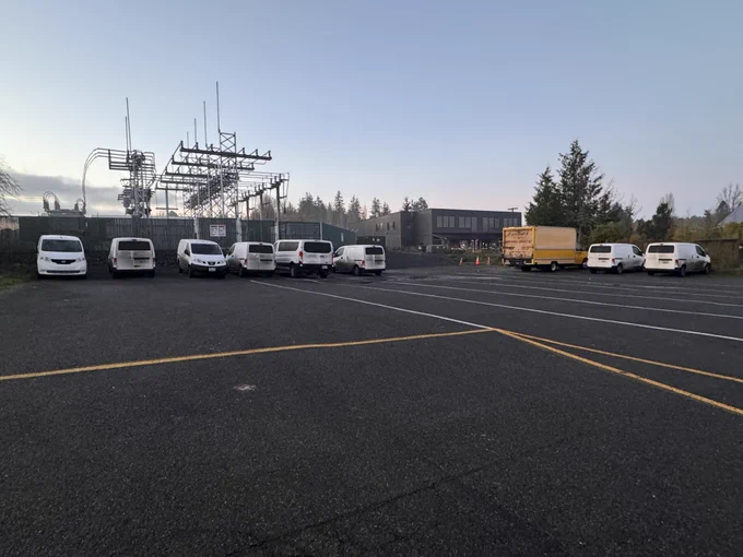 A parking lot with eleven white vans, a yellow box truck, and an electrical substation in the background.