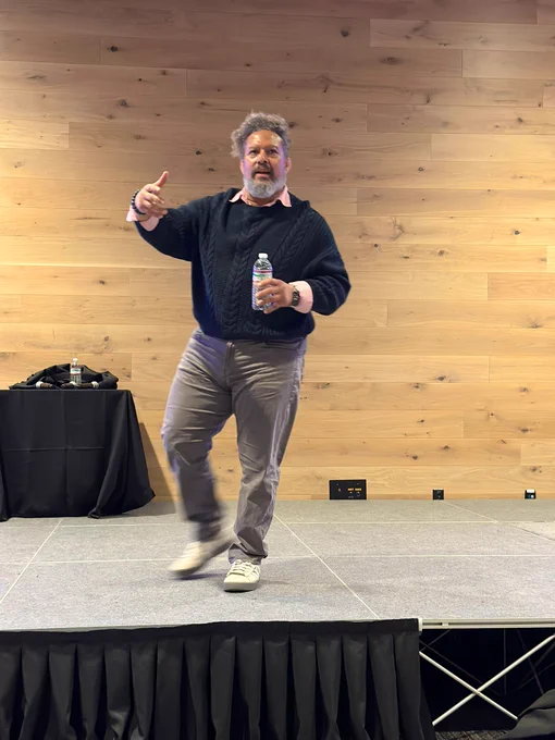 Tim Banks wearing a black sweat let and pink dress shirt underneath, grey pants, on stage delivering Keynote