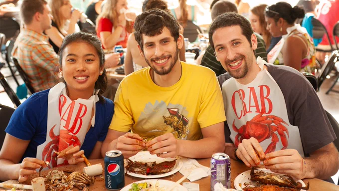 Three students sit together, smiling at the camera. Each student has a crab on their plate in front of them, their fingers covered in old bay seasoning. The two students on the end wear plastic bibs with a drawn image of a crab and the word “CRAB” above it. 