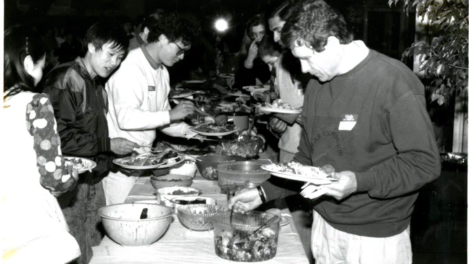 A black and white photograph of several students lined up on either side of a long table, with bowls on it lined up potluck style. Each student fills their plates as they move down the line. 