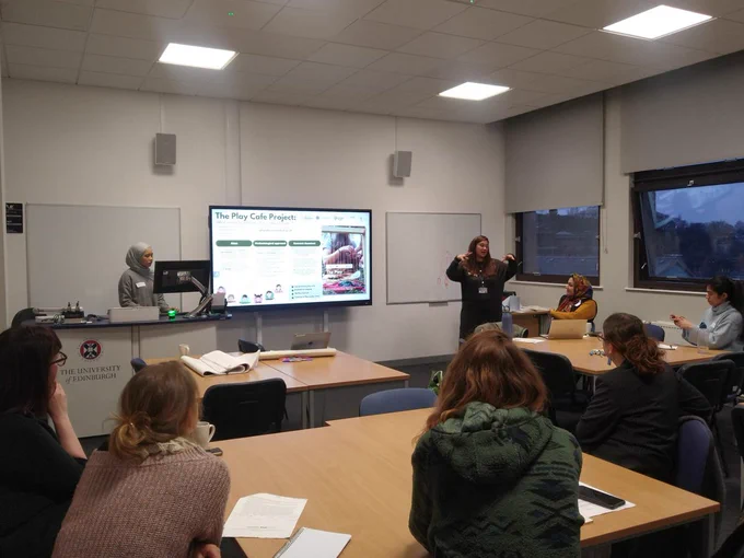Dr Kristina Konstantoni is standing at the front of the room facing workshop attendees and presenting on the Play Cafe project