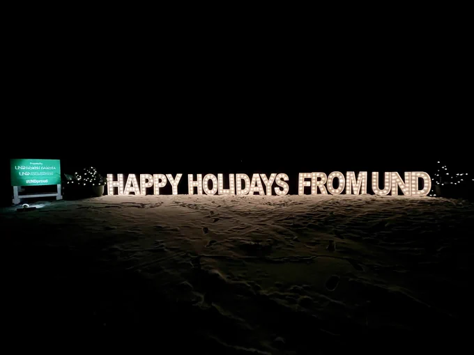 Nighttime view of a snowy landscape illuminated by a lit sign that reads "HAPPY HOLIDAYS FROM UND," with a smaller sign referencing the University of North Dakota nearby.
