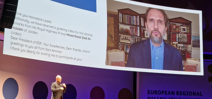 Projected image of His Royal Highness Prince Mired Raad Zeid Al-Hussein of Jordan, giving recorded remarks at the end of the European Regional Disability Summit in Berlin. His image is projected on a large screen on the stage, with a sign language interpreter also visible.