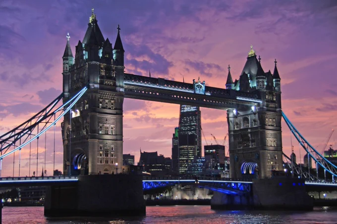 Twilight view of Tower Bridge in London, illuminated with blue lights, against a vibrant sunset sky.