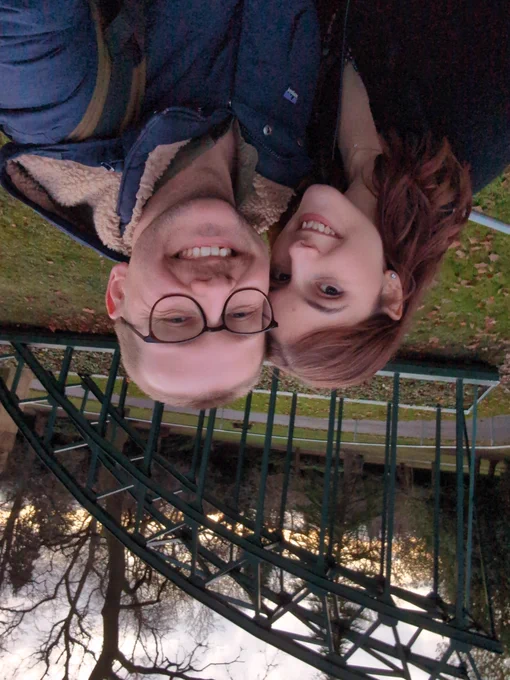 The Tweeter and his very beautiful girlfriend in front of a model of a bridge in Durham. They're very happy, with big smiles. The sun is setting in the background, visible through the gaps in bare winter trees. 