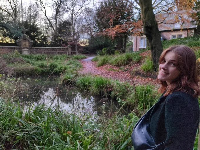 A nice autumnal / early winter scene: a little pond, the last of the red leaves from a tree covering the path at its edge. The sun is setting in the background, the same scene as the last photo. The Tweeter's girlfriend is at the right of the photo smiling; radiant. Her hair matches the leaves.