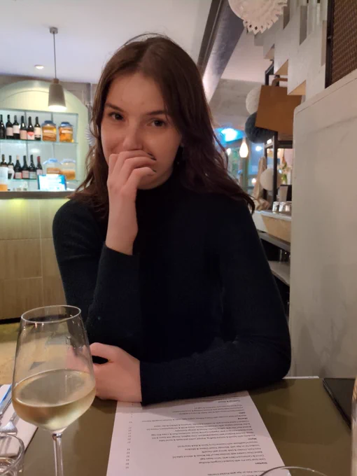 The Tweeter's girlfriend in an indoor scene, a more dimly lit restaurant. She is covering a smile with her hand. She has a glass of wine.