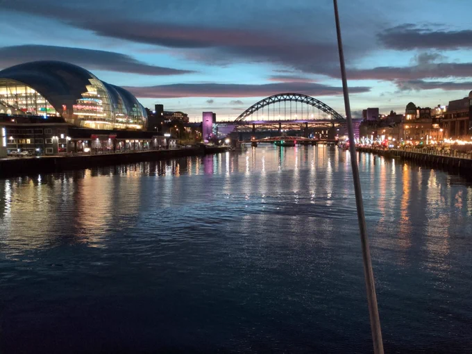The Tyne in Newcastle at late sunset. There are a few diffuse grey clouds, the river reflects the light from the city on its banks and the last of the light in the sky, rippled with moving water. There is a bridge in the distance.
