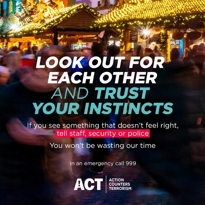 The foreground shows blurred motion of people walking in a crowded area, suggesting a public place like a market or festival. 

In the mid-ground, large, bold text reads "LOOK OUT FOR EACH OTHER AND TRUST YOUR INSTINCTS". 

Beneath this, smaller text provides instructions: "If you see something that doesn't feel right, tell staff, security or police. You won't be wasting our time". 

Further down, smaller text states "In an emergency call 999". 

The background shows festive lights and structures, implying a festive or market setting, with blurred figures of people. The overall tone is serious but concise, focusing on safety and urgency.