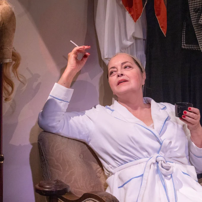 Greta Scacchi wearing a white dressing gown leans back in an armchair, she holds a cigarette in one hand. 