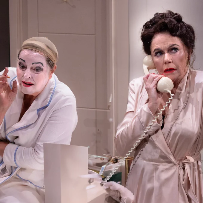 Greta Scacchi with white make-up and red lipstick leans towards Felicity Dean, who is dressed in a pink robe and talks on the phone. 