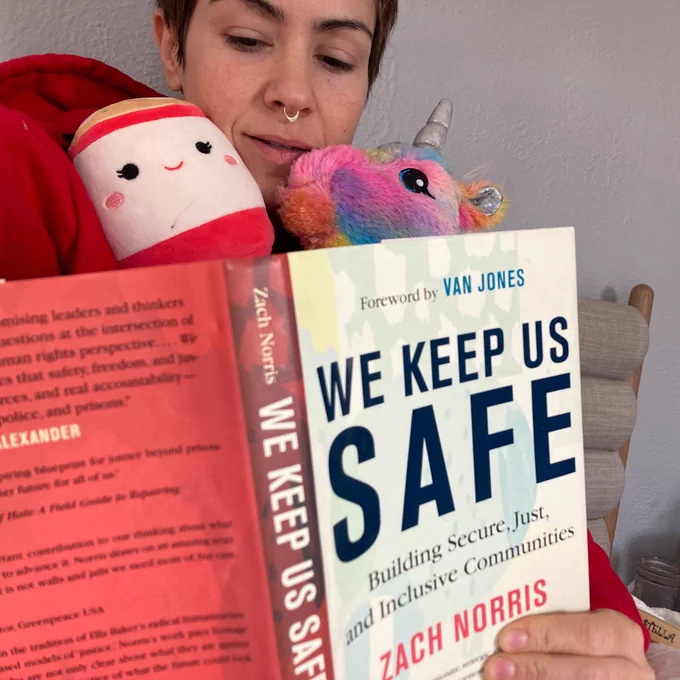 Book cover of We Keep Us Safe by Zach Norris takes up most of photo. My hand holding it, top of my head behind it, mouth open, with rainbow unicorn and ramen squishmallow stuffies in arms.