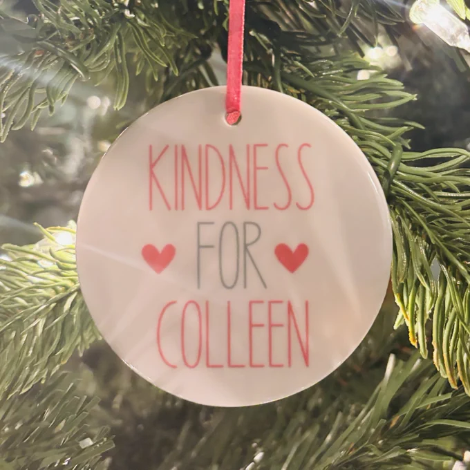 White round ornament that says KINDNESS FOR COLLEEN hanging on a gree tree.
