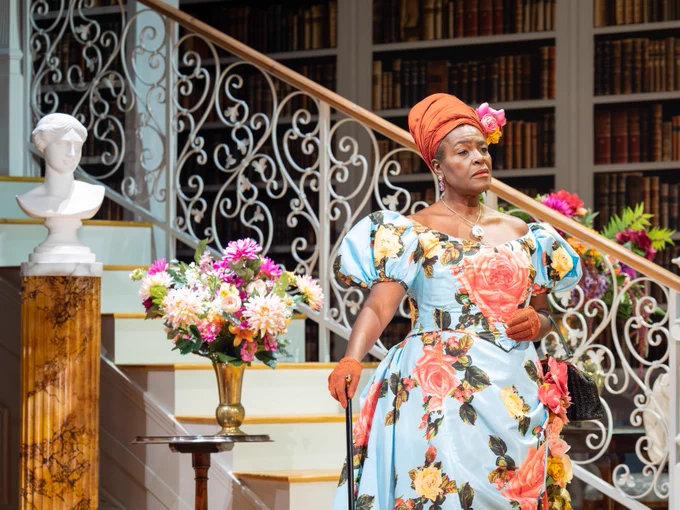 A woman wearing a bold floral dress and headpiece stands against a lavish room backdrop of a large staircase, bouquet of flowers and a marble bust.
