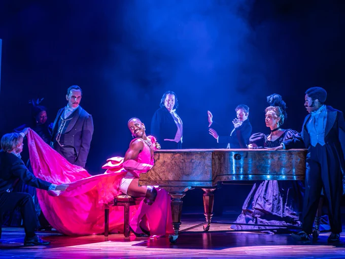A man wearing a pink dress plays a piano on-stage as people wearing suits surround them.