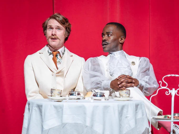 Two men wearing white suits sit as a table with afternoon tea.