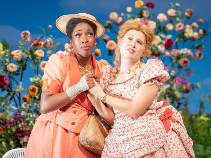 Two women wearing floral dresses and one wearing a boater hat hold hands in a garden filled with flowers. One clutches a handbag.