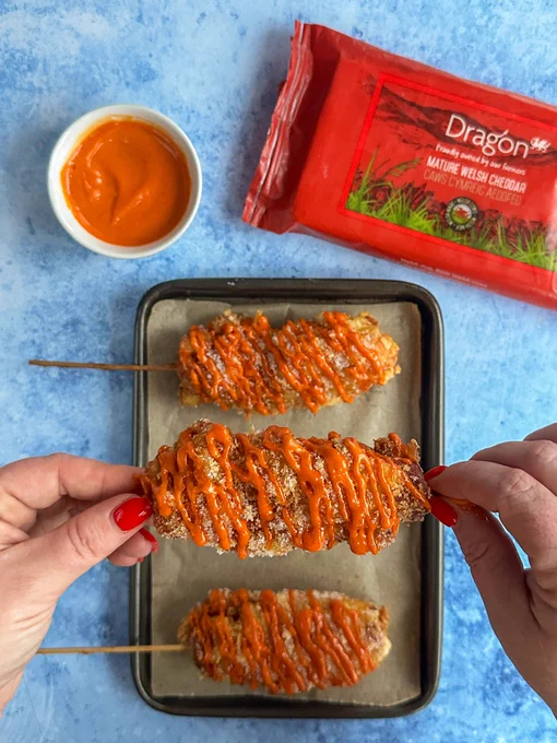 Korean corn dogs on a baking tray, sprinkled with sugar and drizzled with gochujang mayo. Next to the tray, there is a small bowl of mayo and a packet of Dragon Wales Mature Welsh Cheddar. Kacie is holding one of the corn dogs up to the camera