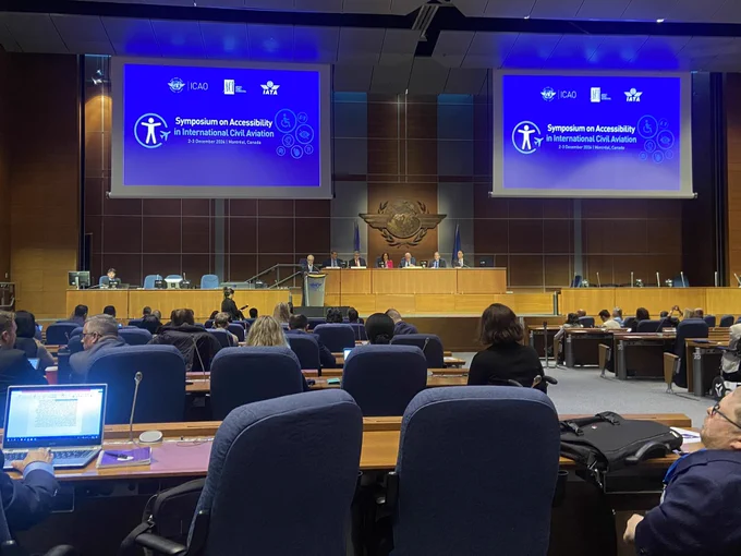 A view from the audience with “Symposium on Accessibility in International Civil Aviation” written on the screens on either side of the stage.