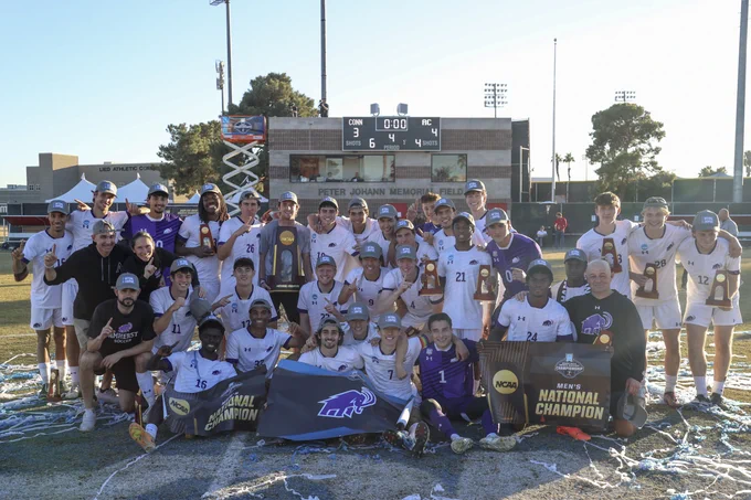 Group photo of the Amherst College men's soccer team celebrating their NCAA national championship victory on a sunlit field.
