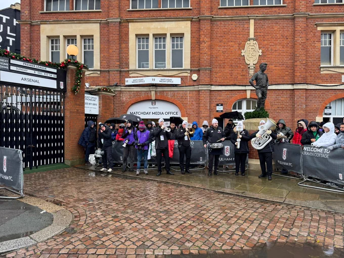 Photo of outside craven cottage by the Johnny Haynes Stand with a band playing and fans waiting for the club coaches carrying the players to arrive. Kickoff soon!